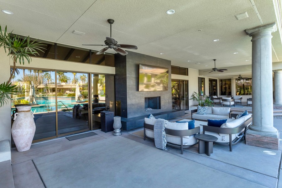 Luxurious covered patio featuring a modern outdoor fireplace with a mounted TV, stylish seating area, ceiling fans, and a view of a backyard pool surrounded by palm trees.